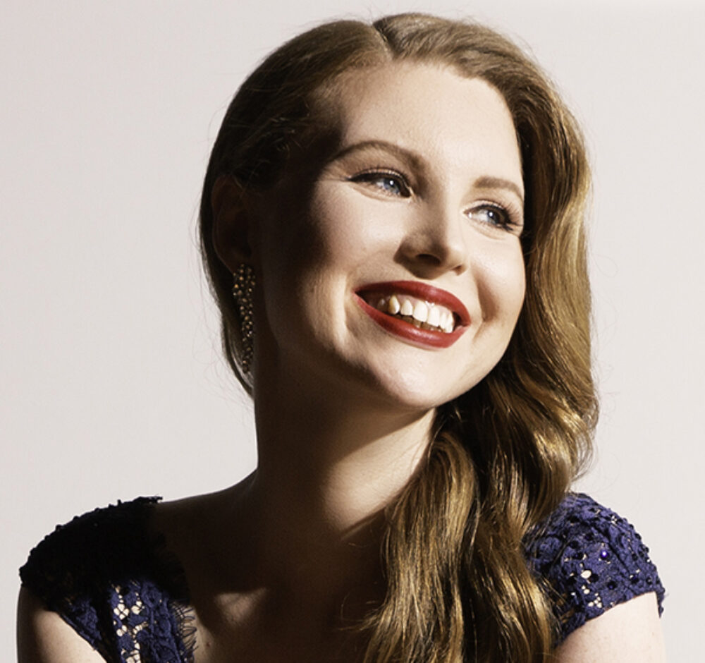 Carolyn smiles brightly with her head tipped to the right of the camera. She has red hair and bright red lipstick.