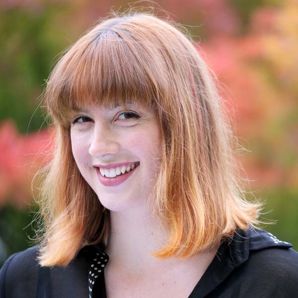 Gina smiles brightly as she is turned slightly away from the camera, but looking directly at the viewer. She has shoulder length ginger hair with bangs and wears red lipstick.