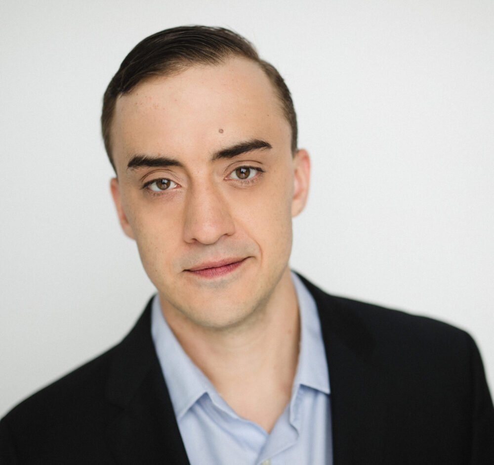 Geoffrey looks into the camera, short dark hair slicked back, wearing a black suit jacket and blue button up.