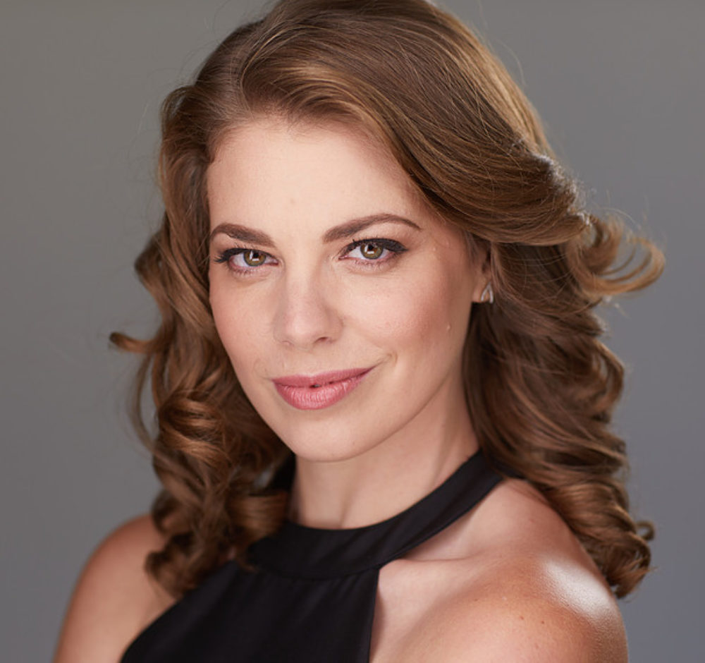 Marjorie smiles mischievously, her caramel brown hair curled and swept back behind one shoulder. She has hazel eyes and wears a black halter neck shirt.