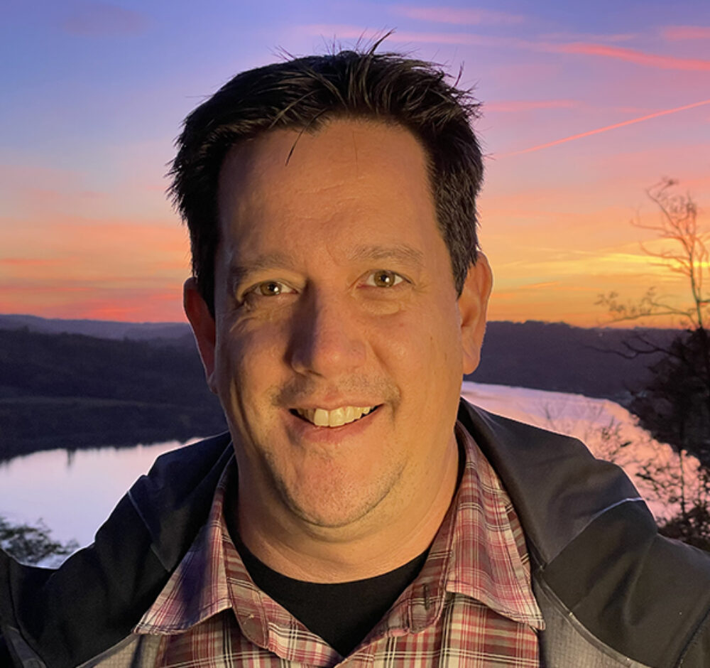 Carey Newman smiles at the camera, wearing a light red and white plaid shirt and a green jacket. There is a lake and a purple, orange sunset behind him.