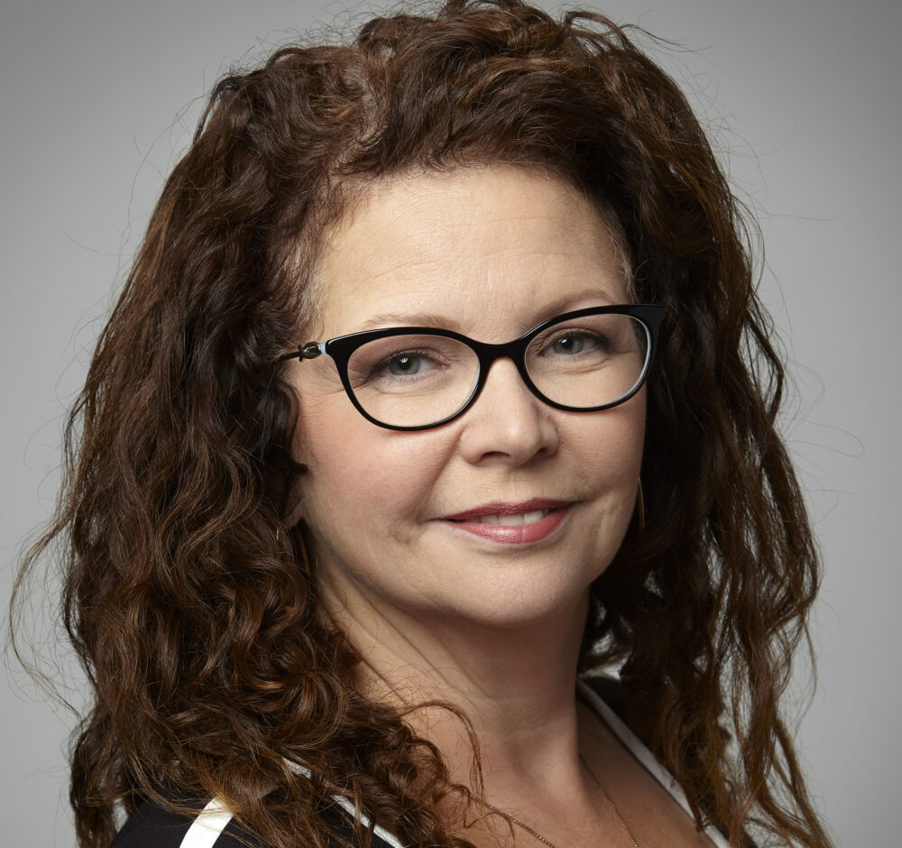 Deneen smiles, her body turned slightly away from the camera. She has curly red-brown hair and wears cat-eye glasses.