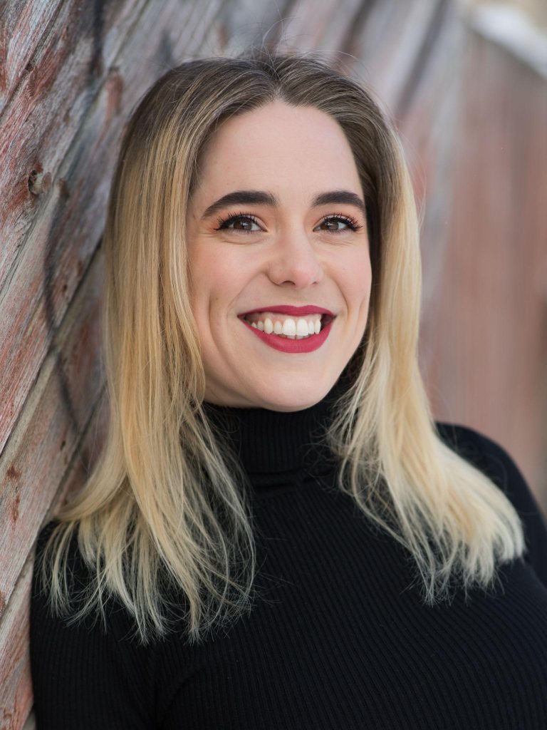 A photo of soprano Chelsea Kutyn from her shoulders up. She is smiling and has shoulder length blond hair with dark roots.
