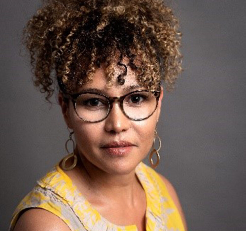Paulina looks directly into the camera, her eyes framed by glasses. Her curly hair falls over her forehead and she is wearing a yellow pattered shirt.