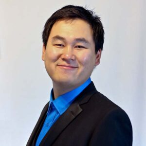Dressed in a black suit jacket and royal blue button up shirt, Alex smiles softly at the camera, with his body turned slightly to the side. Alex has short black hair.