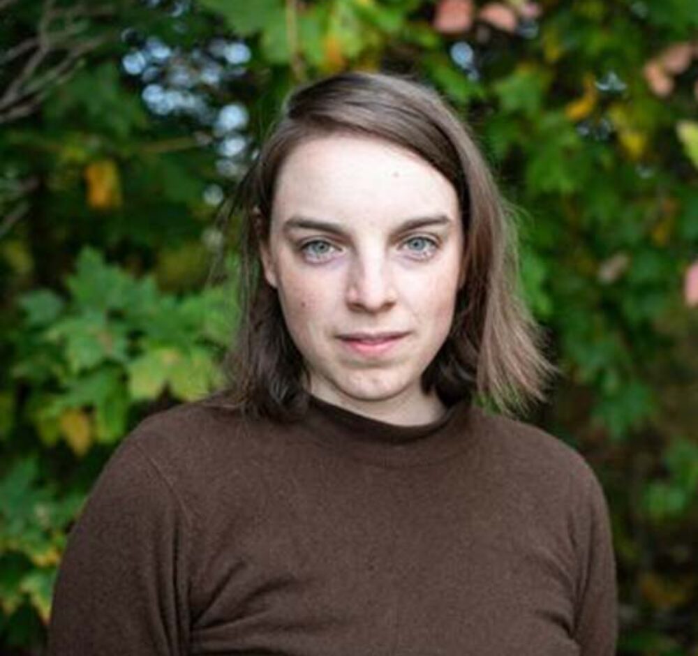 Rebecca stares down the camera, wearing a brown long sleeve shirt and standing in front of a bush. She has shoulder length straight brown hair that is tucked behind her ear on one side.