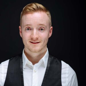 Tim is dressed in a white button up shirt with a black vest overtop. Smiling in front of a black background, Tim's ginger hair and blue eyes stand out as he smiles at the camera.