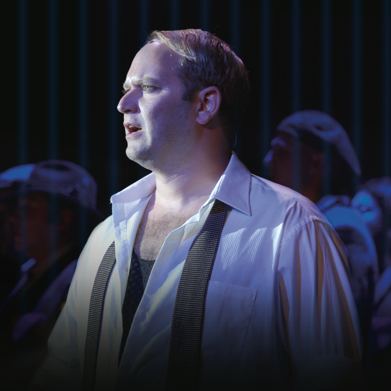 Manfred is dressed in a white shirt that is unbuttoned about halfway and an undone tie. He looks to the side with a look of anguish on his face. men standing behind him, dimly lit.