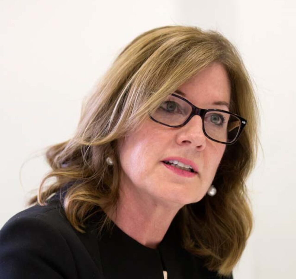 Elizabeth looks out past the camera with a thoughtful look on her face, wearing a black suit jacket and glasses. She has light brown hair that is slightly curled and sits at her shoulders.