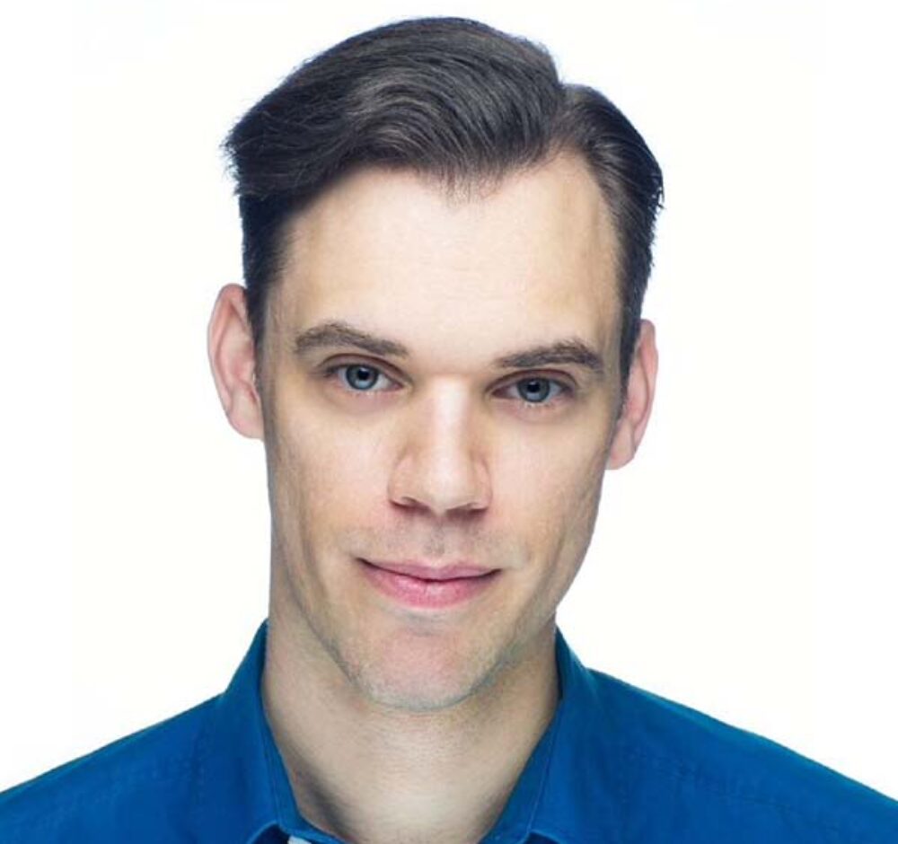 Geoff smiles softly at the camera as he stands against a white background. His brown hair is smoothly swept to the side and he wears a blue button up.