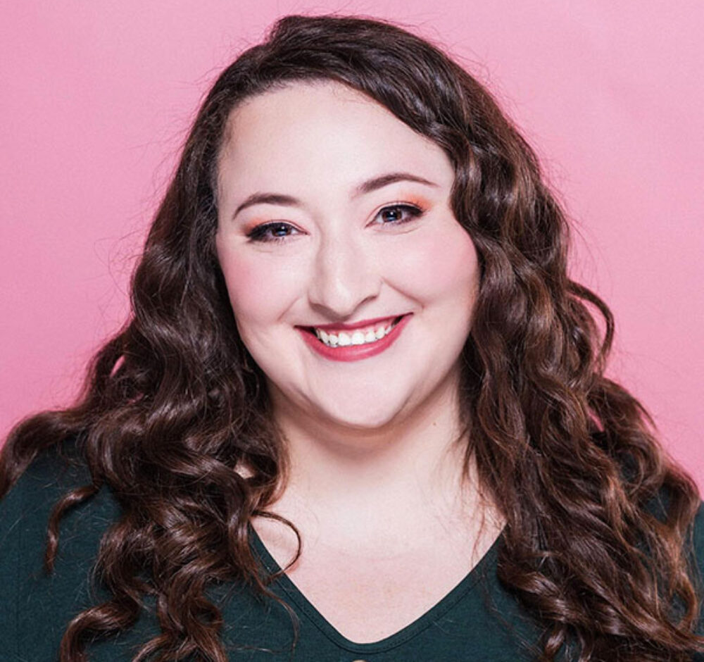 Jaclyn smiles directly at the camera, with pink lipstick and brown curly hair. She is against a light pink background and is wearing a green shirt