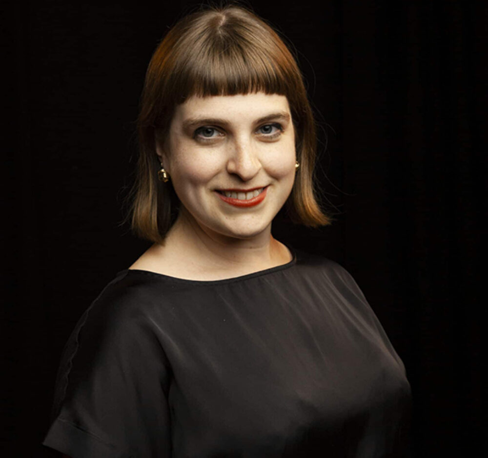 Juliane smiles at the camera, wearing a black silk shirt against a black background. She has shoulder-length straight brown hair and bangs, and wears red lipstick.