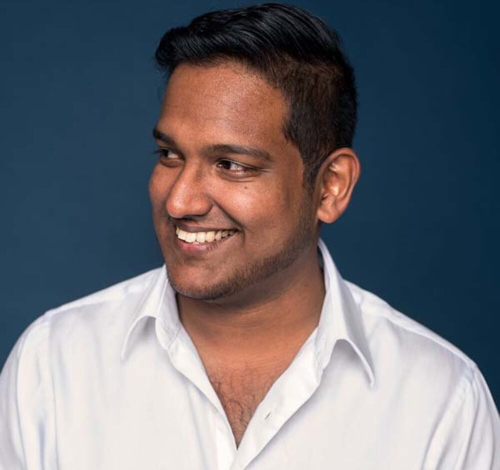 Asitha smiles warmly, looking to the left of the camera. He has close cropped black hair that is longer on top and is wearing a white button up.