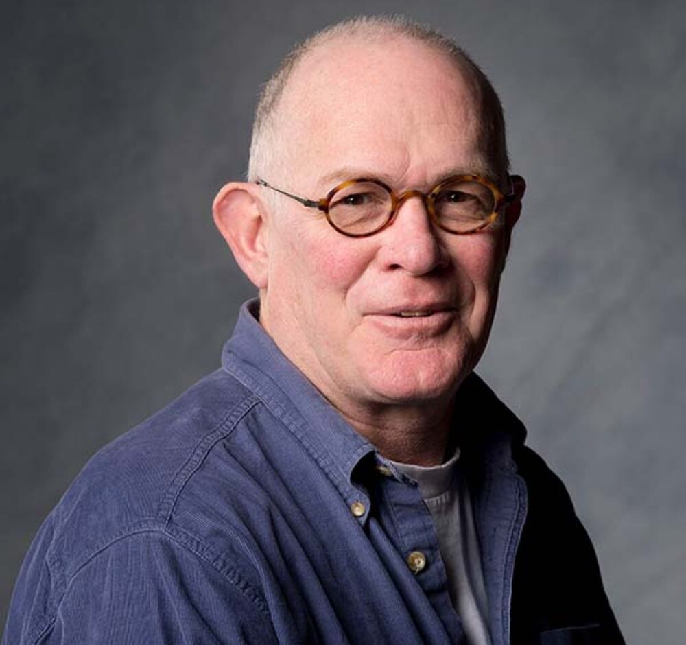 Peter smiles softly in a blue buttton up and small brown round framed glasses.