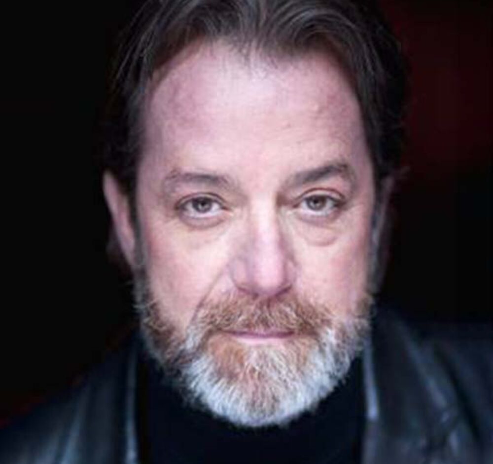 C David looks directly at the viewer, wearing a leather jacket an black shirt against a black bavkground. He has black hair and a gray beard.