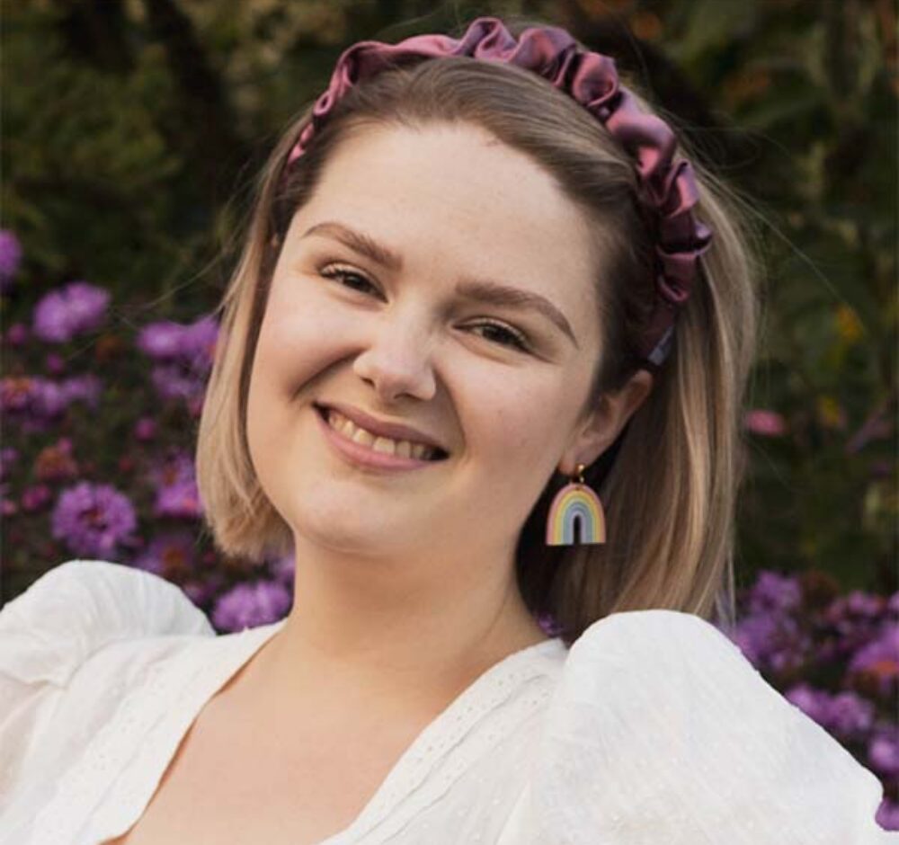 Anna smiles brightly at the camera, shoulder length blonde hair held back with a purple headband. She wears a white shirt with puffy sleeves and stands in front of purple flowers.