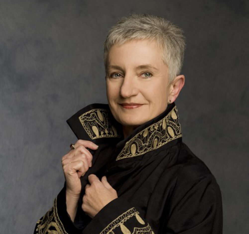 Christina stares boldly at the camera, holding onto the collar of her jacket with both hands. It has a thick gold trim. She wears red lipstick and has gray hair.