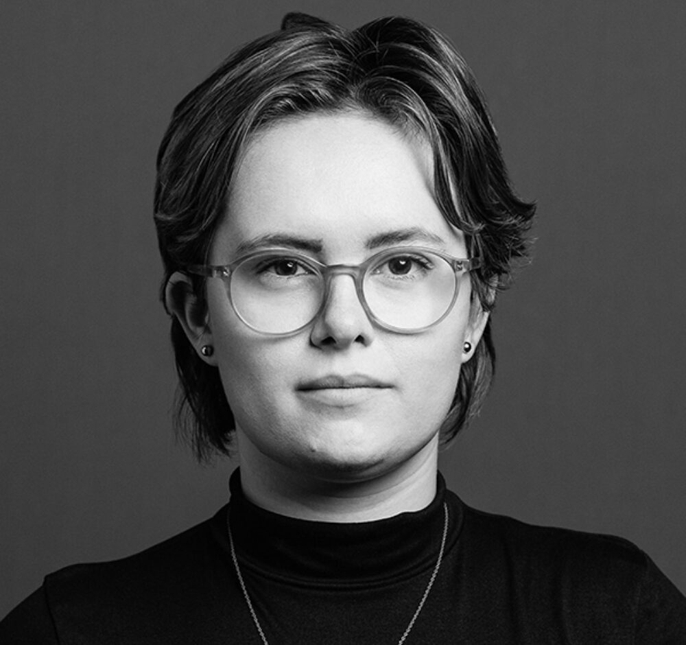 Jamie stares into the camera, wearing a black turtleneck and square framed glasses. She has short hair in this black and white photo.