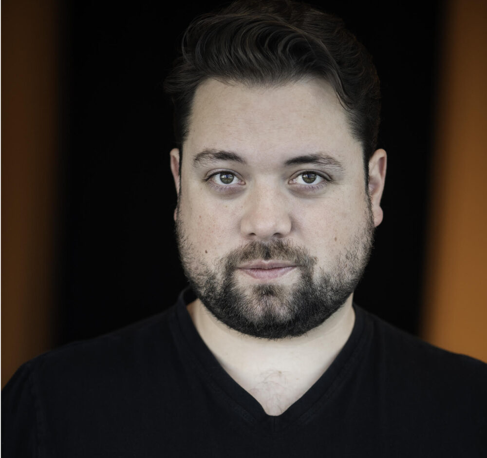 Geoffroy has dark brown hair and a beard. He wears a black shirt.