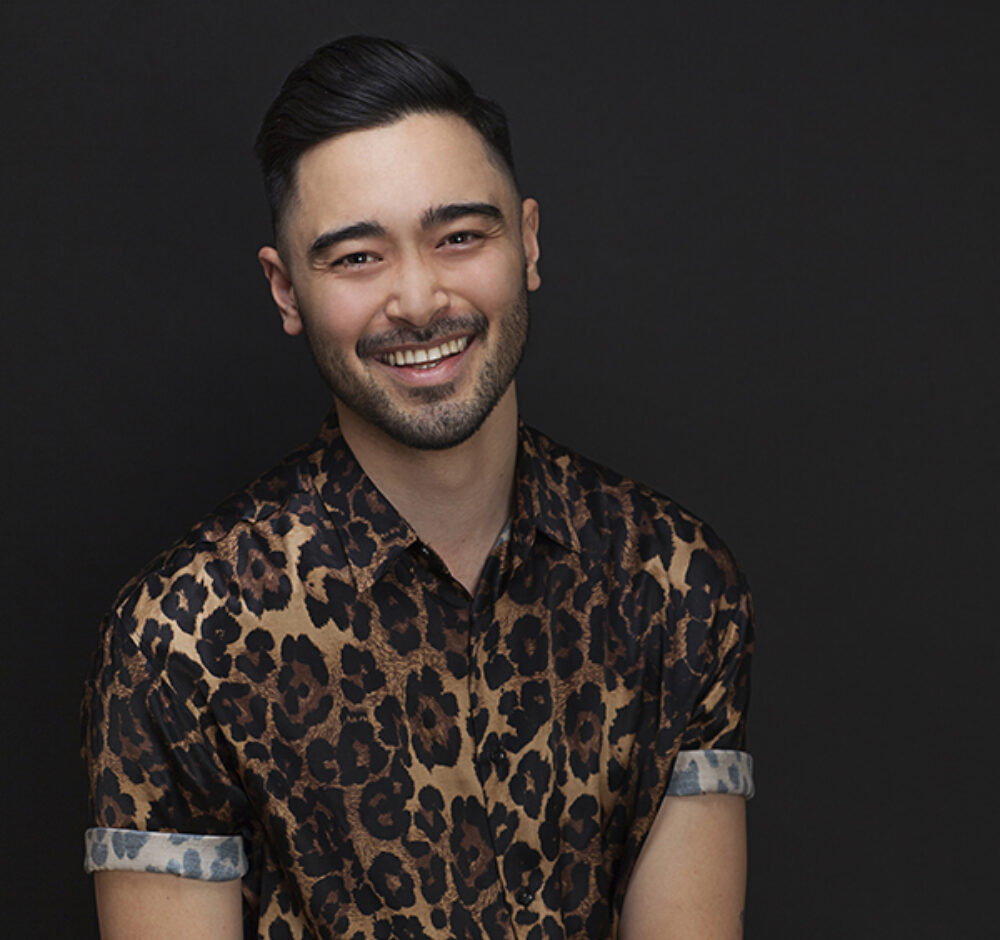 Luka smiles brightly at the camera, a dark blue background behind him. He had a 5'o'clock shadow and black hair. He is wearing a cheetah print, short sleeve button up shirt.