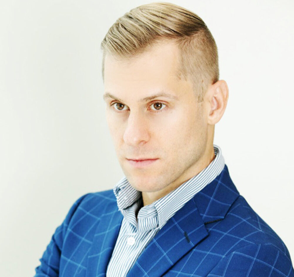 Adrian looks to the left of the camera with a bold stare. He wears a light blue button up shirt with a royal blue suit jacket that has a thin lined grid pattern. He has short blond hair that is slightly longer on top and brown eyes.