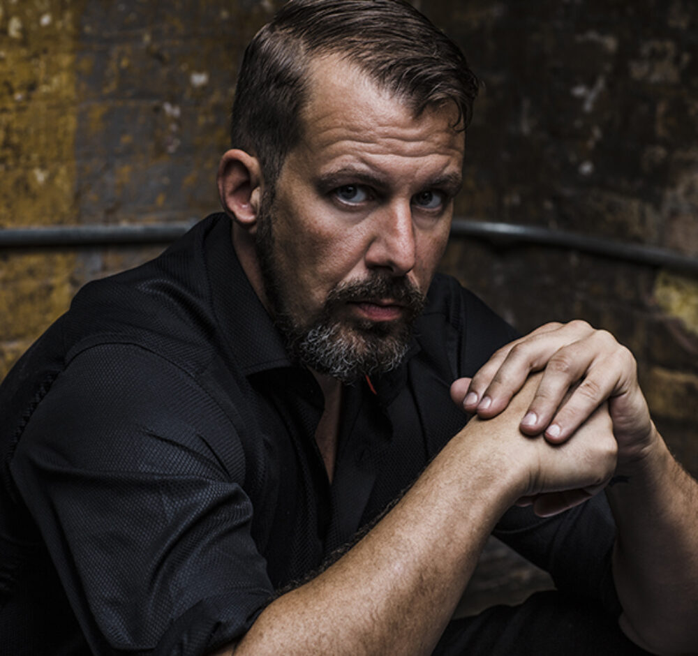 Kyle stares deeply into the camera, his body turned parallel to it with his elbows resting on his knees and one hand on top of the other. He wears a black button up shirt with the sleeves rolled above his elbows. He has dark hair and a beard.