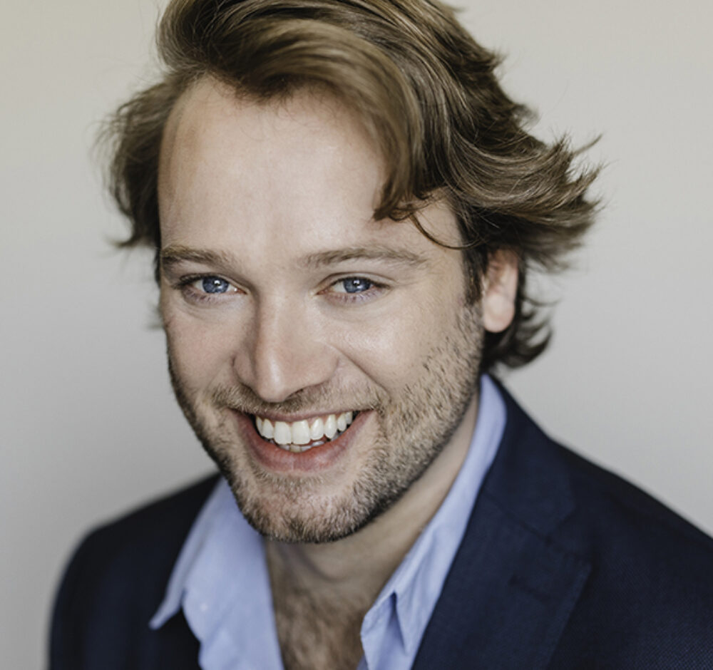 Nathan smiles brightly, his dirty blonde hair swept around his face, going in multiple directions. He wears a light blue button up and dark blue suit jacket.