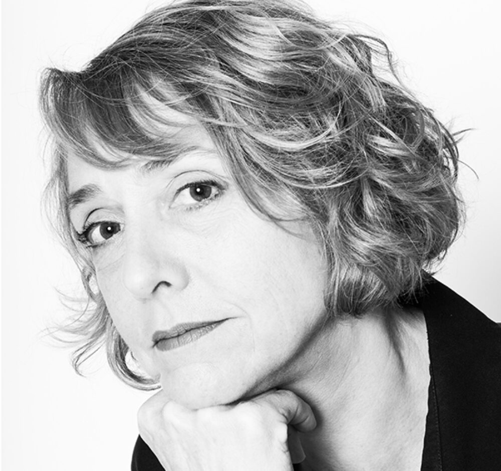 Chantal rests her head on one hand, looking at the camera. Her curly hair is cut to chin-length and she has bangs resting on her forehead in this black and white image.