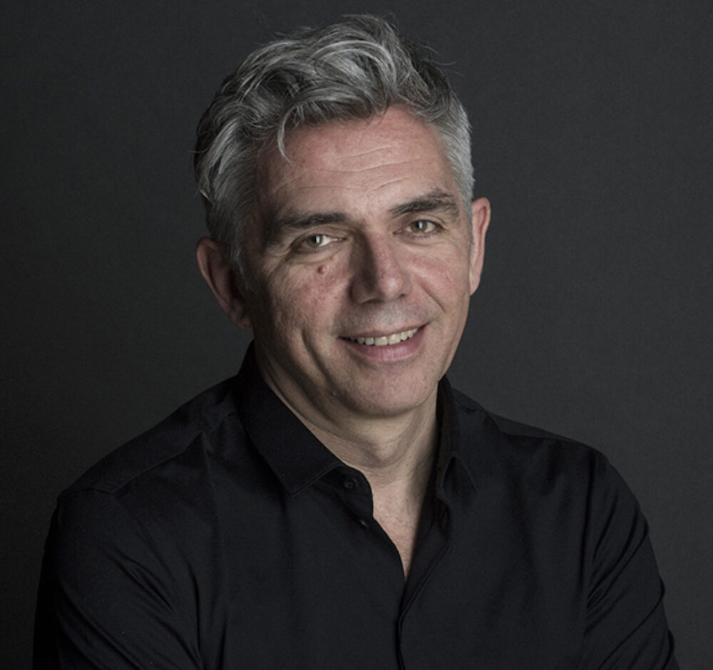 Laurent smiles, wearing a black button up. He has short gray hair and hazel eyes.