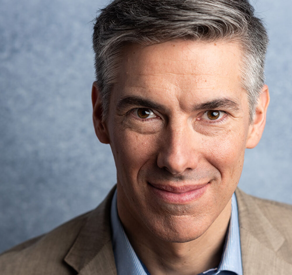 Phillip smiles slightly, wearing a blue button up and tan shirt. He has short grey hair.
